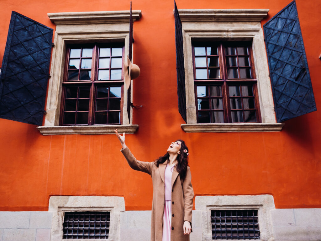 A person in a long coat is standing in front of an orange building with two windows. The person is looking up and reaching toward a hat that appears to be falling or floating above them.