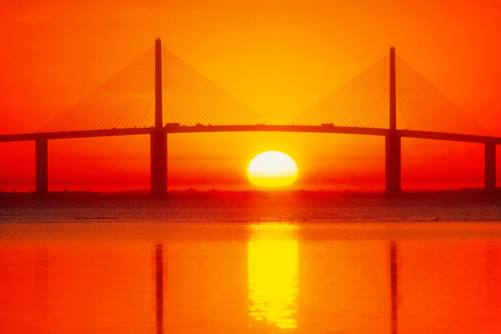 Un puente con altas torres de apoyo cruza una masa de agua al atardecer, silueteado contra un cielo naranja y rojo con el sol parcialmente bajo el horizonte, simbolizando el viaje de formación de la próxima generación de expertos chinos.