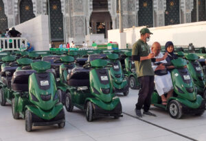 A person assists an elderly man and woman on a motorized scooter amid a row of similar scooters, set against the backdrop of a large, ornate building.
