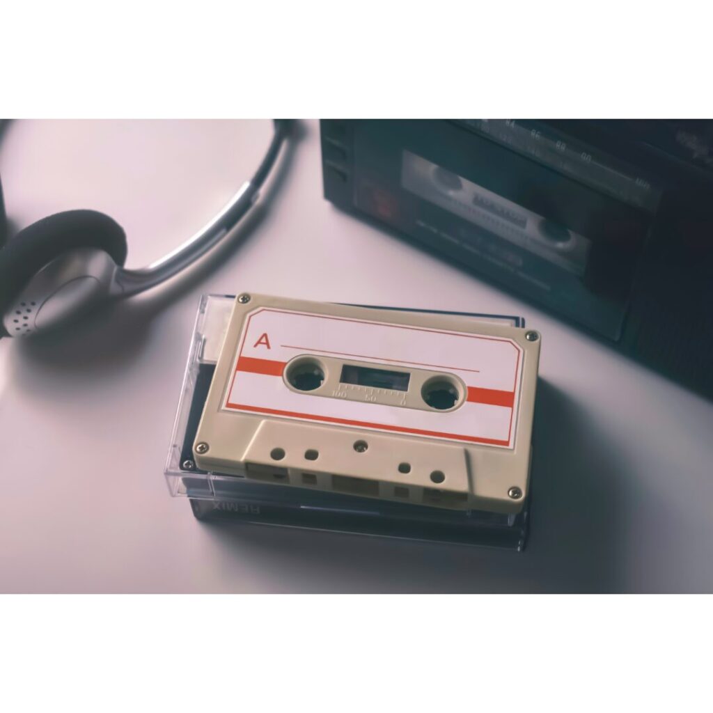 A cassette tape labeled "A" is placed on a clear plastic case next to a pair of over-ear headphones and a cassette player on a white surface.