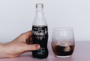 A hand holding a coca-cola bottle next to a glass half-filled with cola on a light pink surface against a plain background.