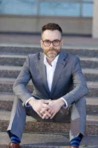 Vince Sliwoski, a man with a beard and glasses, sits on stone steps wearing a gray suit and blue striped socks, his hands clasped in quiet contemplation.