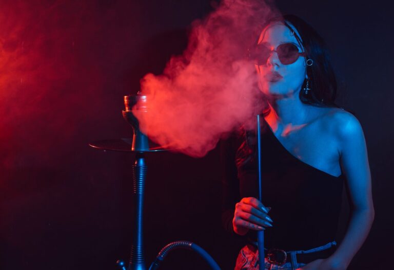 Person in sunglasses blowing smoke while holding a hookah pipe, surrounded by red and blue lighting in a dark room.