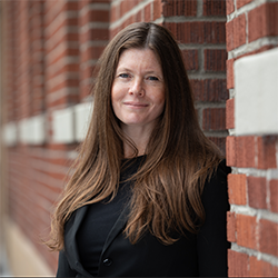 Deb Fernando, with long brown hair in a black outfit, stands against a brick wall, smiling.