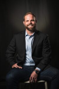 Jared Schwass, a man with a beard in a dark blazer and blue shirt, sits on a stool, smiling, with one hand on his knee against a dark background.