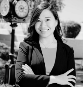 Woman in a blazer stands outdoors with arms crossed, smiling at the camera. A decorative clock and trees are visible in the background. Image is in black and white.