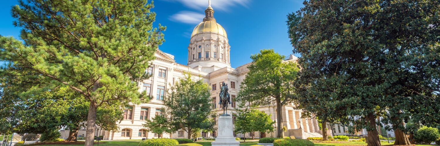 A large government building with a golden dome, surrounded by trees and a statue in front, under a clear blue sky.