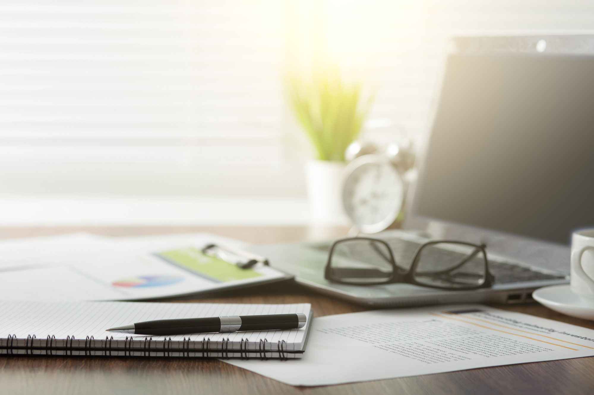 A workspace with a laptop, glasses, a pen on a notebook, documents, a clipboard, a potted plant, and an alarm clock, all placed on a wooden desk. Sunlight filters through blinds in the background.