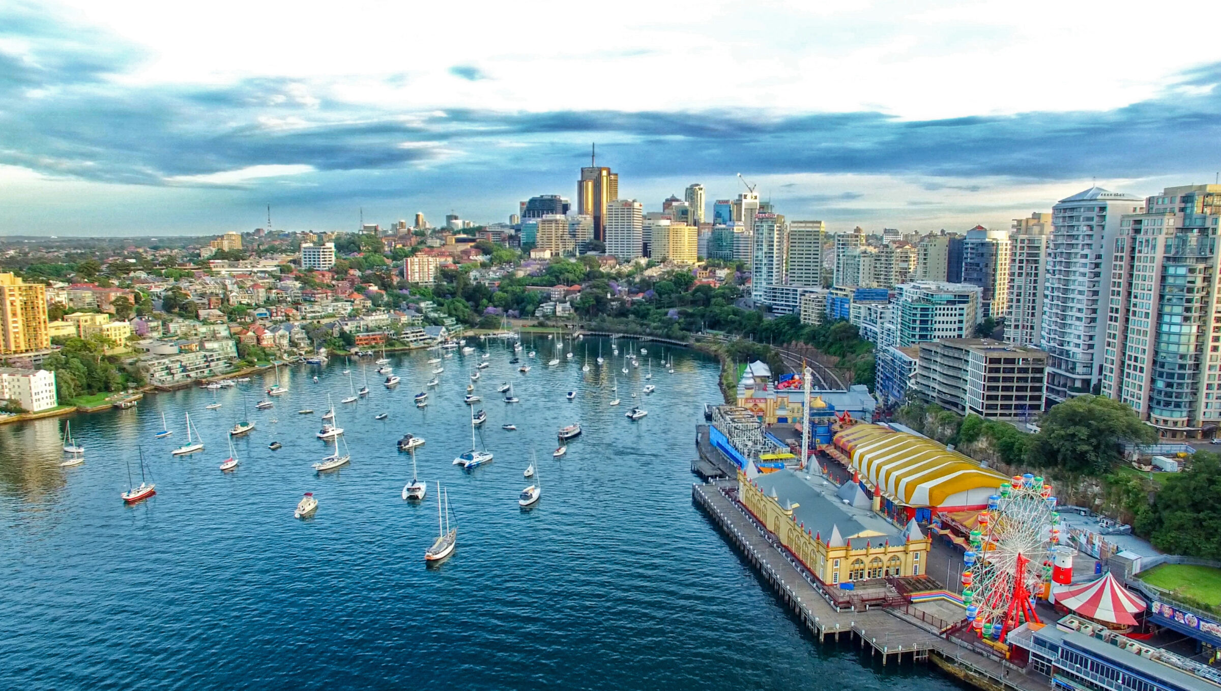 Aerial view of a harbor filled with boats next to a colorful dockside amusement park and surrounded by city buildings and green spaces on a partly cloudy day.