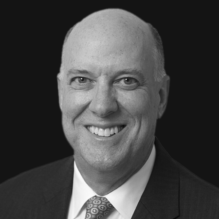 Black and white headshot of Timothy Alger, a bald man in a suit and tie, smiling at the camera against a dark background.