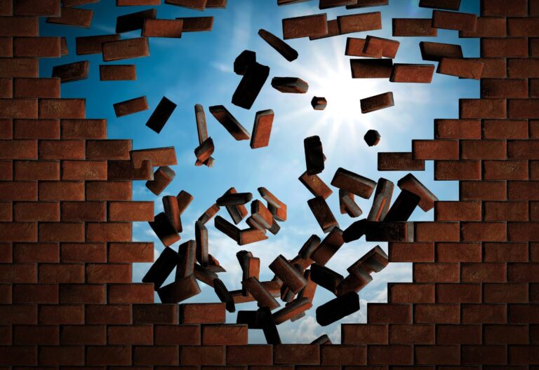 A brick wall with a large hole, showing scattered bricks against a blue sky with clouds and sunlight.