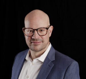 A bald man with glasses wearing a white shirt and blue blazer poses against a solid black background.