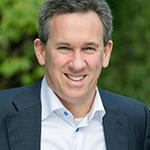 A man in a dark suit and white shirt is smiling outdoors, with a blurred green background.