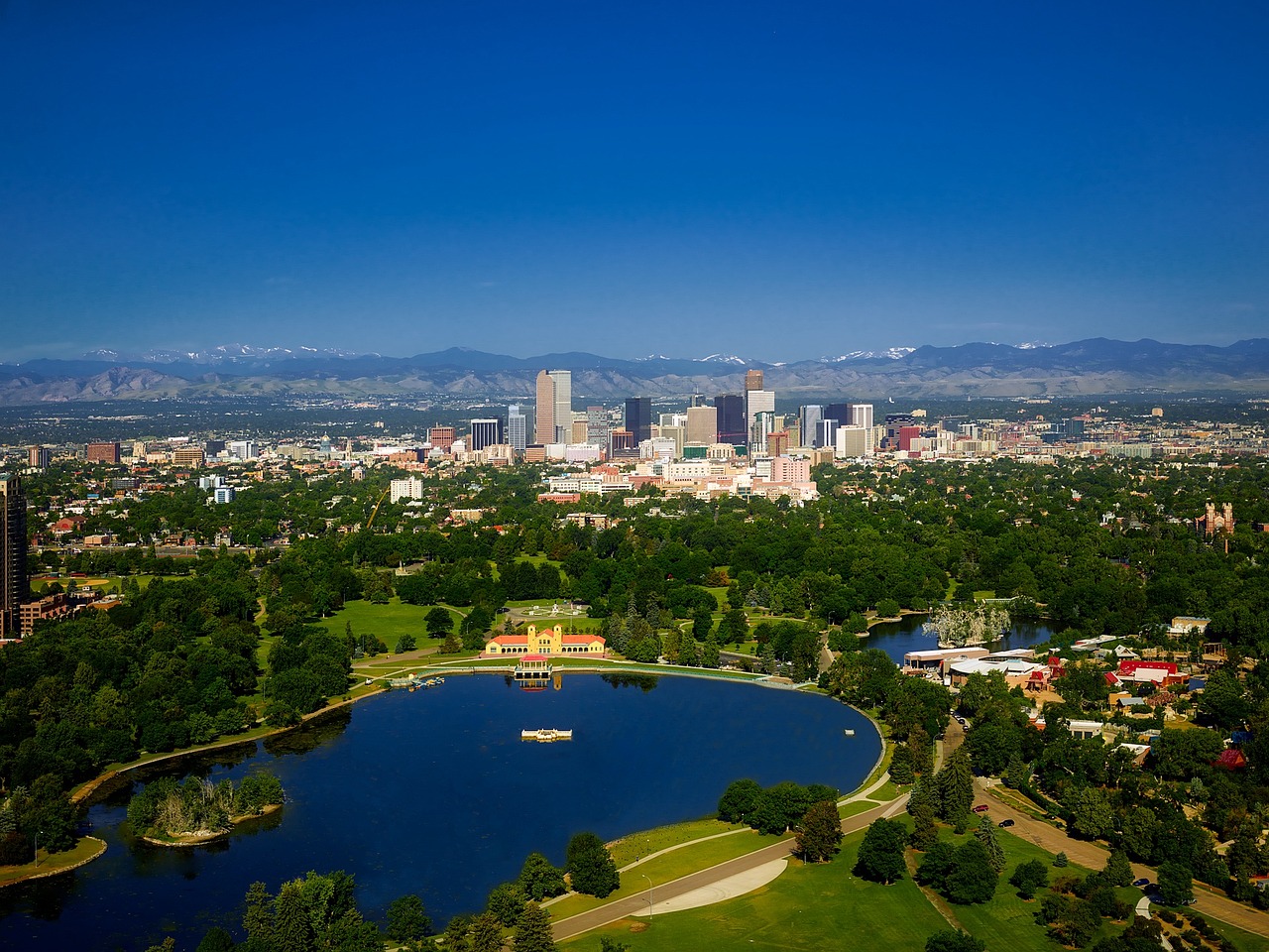 Aerial view of Denver skyline with a large park in the foreground, featuring a lake and trees, with mountains visible in the background under a clear blue sky.