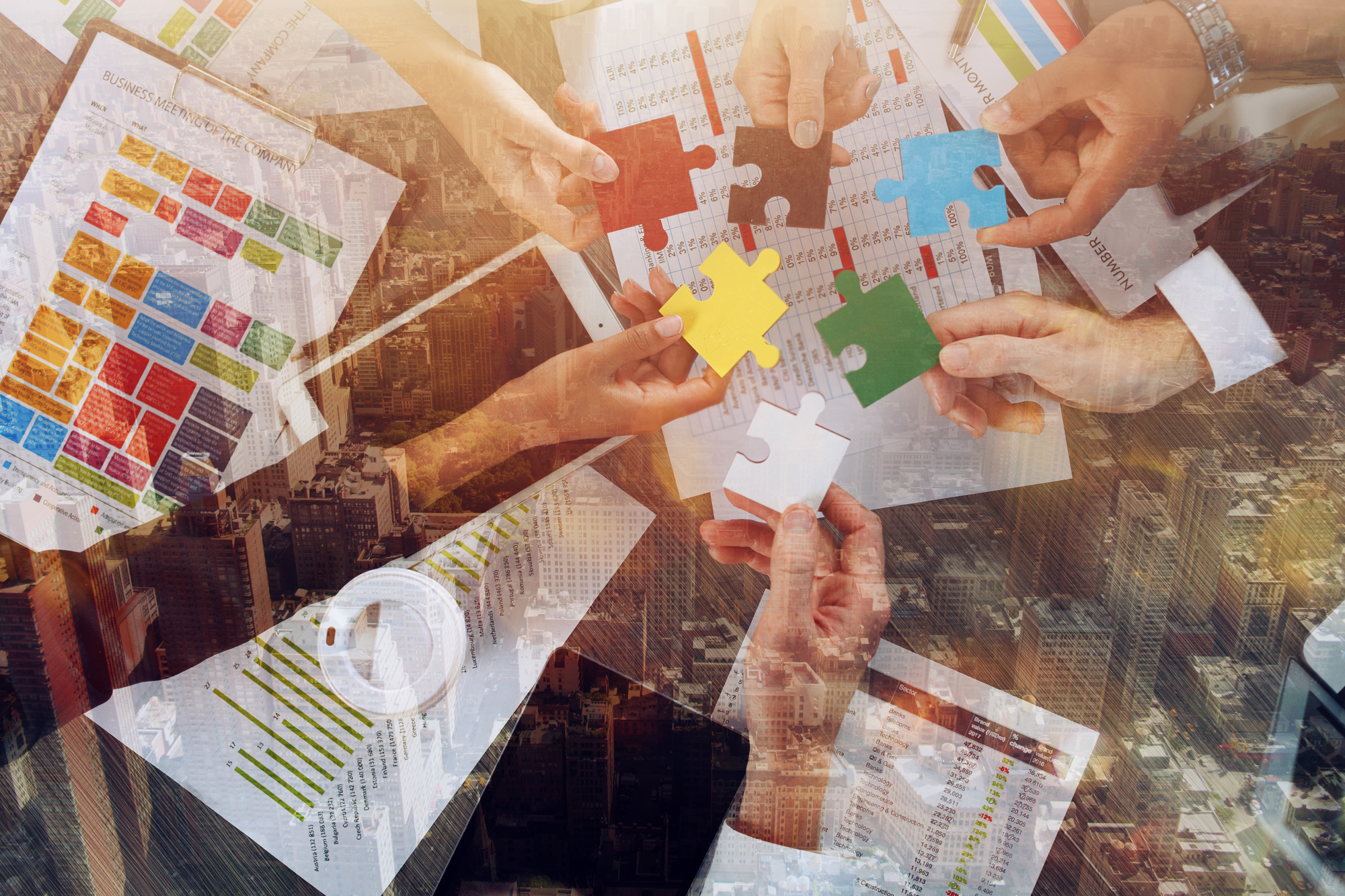 Several hands hold colorful puzzle pieces over documents, symbolizing teamwork and problem-solving, with an overlay of a bustling cityscape.