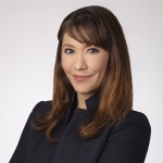 A woman with long brown hair and bangs is wearing a dark blazer and posing with her arms crossed, facing the camera against a plain background.