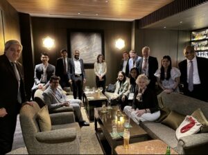 Group of professionals in business attire posing for a photo in a lounge area with sofas and a bar in the background.