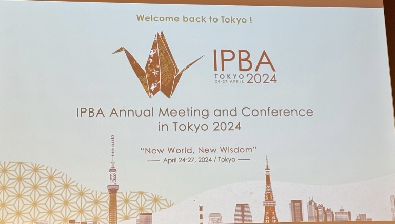 La pantalla de proyección muestra el texto &quot;welcome back to tokyo! ipba annual meeting and conference in tokyo 2024&quot; con un skyline de tokyo ilustrado y la fecha del 24 al 27 de abril de 2024.