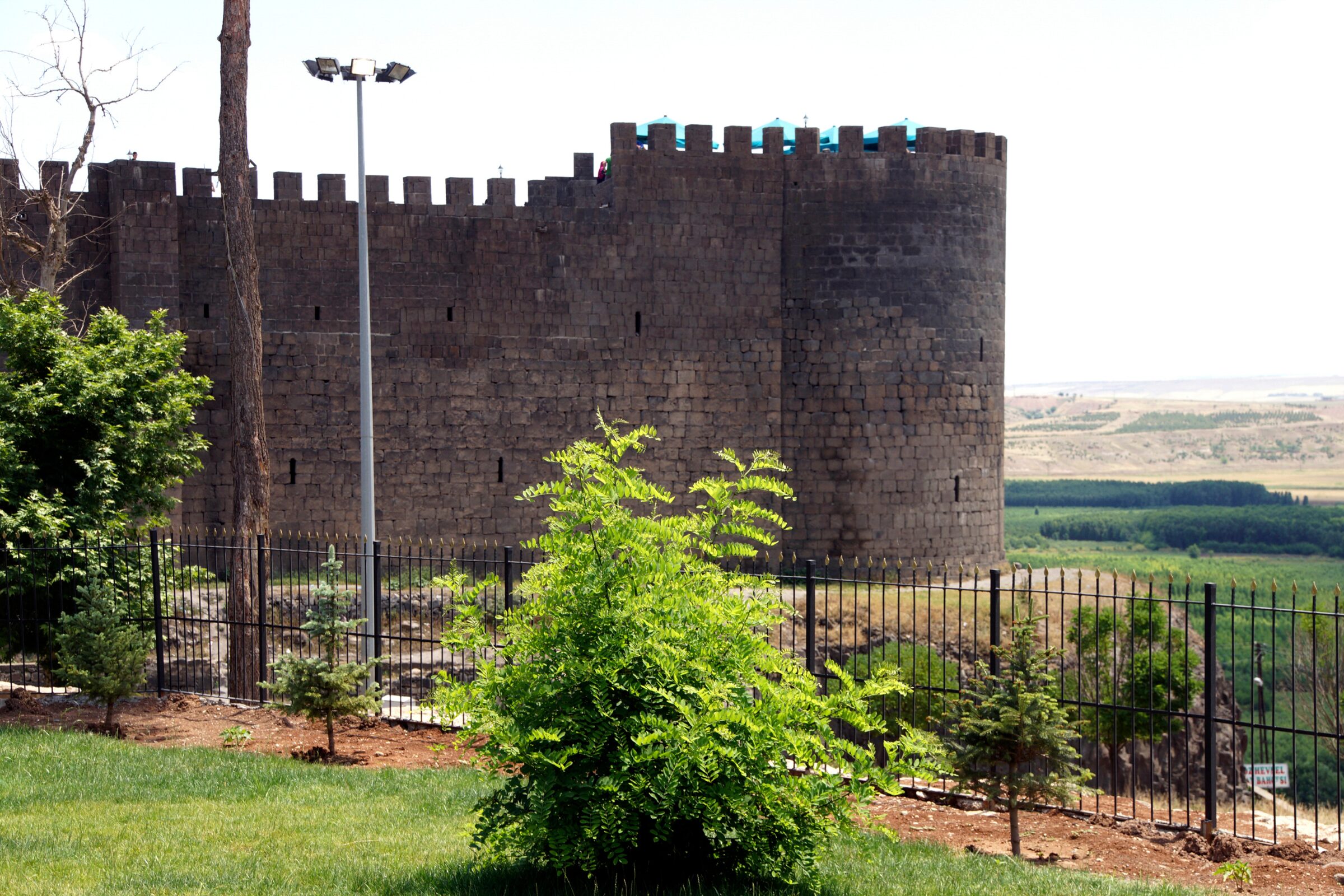 A historic stone fortress wall stands tall behind a black metal fence, surrounded by greenery with trees and shrubs.